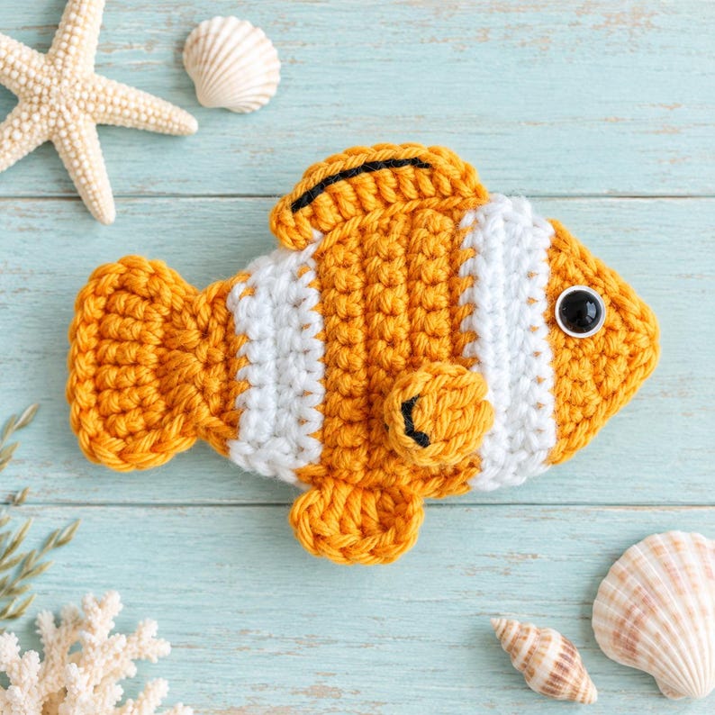 May include: A crocheted clownfish toy featuring vibrant orange and white vertical stripes, a black-rimmed dorsal fin, and a single black plastic eye. The fish is crafted with a textured yarn stitch pattern, showcasing a rounded body, a tail fin, and a small side fin.
