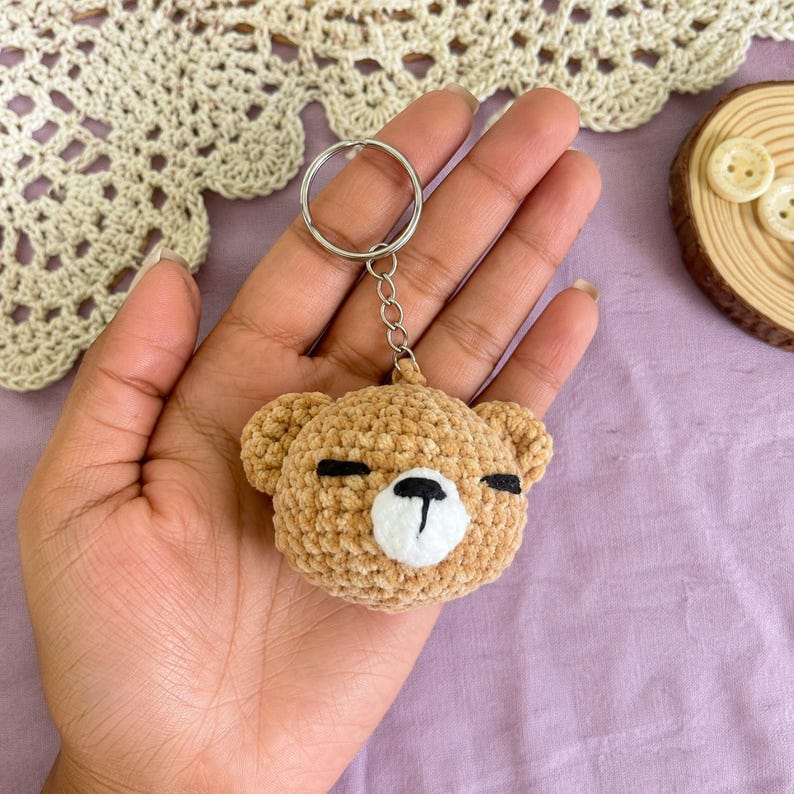 May include: A crocheted tan bear keyring featuring a round head with small ears, a white muzzle, and black embroidered eyes and nose. The bear is attached to a silver-toned metal split ring by a short chain, creating a compact accessory.