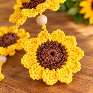 Puede incluir: Una guirnalda de ganchillo con varios adornos en forma de girasol. Cada girasol tiene p&eacute;talos de color amarillo brillante y un centro marr&oacute;n oscuro, conectados por finas hebras de hilo y cuentas de madera redondas, lisas y claras.