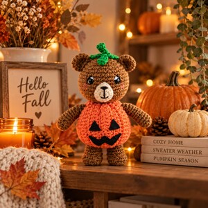May include: A brown crocheted bear plush toy wearing an orange pumpkin-shaped body with a black felt jack-o'-lantern face and a green crocheted stem on its head. The bear has a cream-colored muzzle, black embroidered eyes, and pink cheeks.