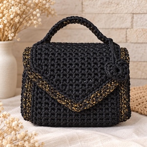 May include: A black crochet handbag featuring a structured envelope flap with gold metallic thread accents along the edges. The bag includes a single top handle and a small, matching black crochet flower charm attached to the side of the handle.