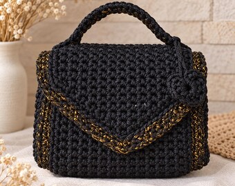 May include: A black crochet handbag featuring a structured envelope flap with gold metallic thread accents along the edges. The bag includes a single top handle and a small, matching black crochet flower charm attached to the side of the handle.
