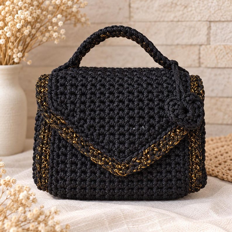 Puede incluir: Un bolso de mano de ganchillo negro con una solapa tipo sobre estructurada y detalles de hilo met&aacute;lico dorado en los bordes. El bolso incluye un asa superior &uacute;nica y un peque&ntilde;o dije de flor de ganchillo negro a juego en el lateral.