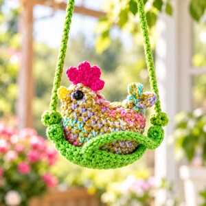 May include: A crocheted chicken ornament featuring a multicolored body with yellow, pink, and blue yarn, a bright pink comb, and a yellow beak. The chicken sits inside a green crocheted swing suspended by two green yarn ropes.
