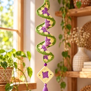 K&ouml;nnte beinhalten: Ein h&auml;ngendes H&auml;kel-Spiralornament mit einem sich wiederholenden Muster aus violetten, cremefarbenen und gr&uuml;nen Granny Squares. Die Spirale ist oben und unten mit violetten Perlen verziert und endet in einem quadratischen Motiv mit Quaste.