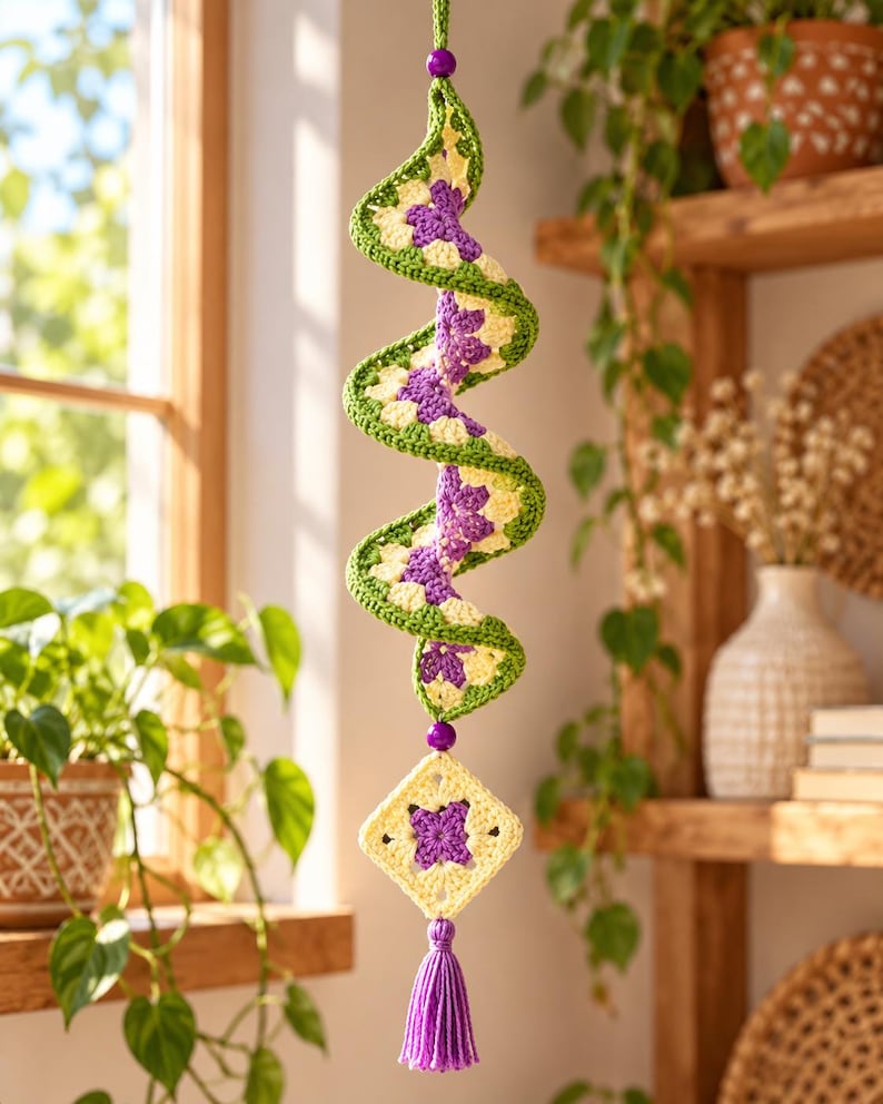 K&ouml;nnte beinhalten: Ein h&auml;ngendes H&auml;kel-Spiralornament mit einem sich wiederholenden Muster aus violetten, cremefarbenen und gr&uuml;nen Granny Squares. Die Spirale ist oben und unten mit violetten Perlen verziert und endet in einem quadratischen Motiv mit Quaste.