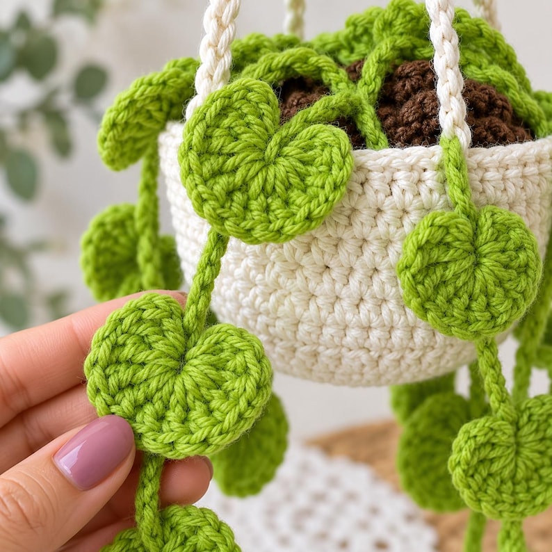 May include: A crocheted hanging plant featuring a cream-colored pot filled with dark brown soil and trailing vines with bright green, heart-shaped leaves. The plant is suspended by thick, cream-colored braided yarn cords.