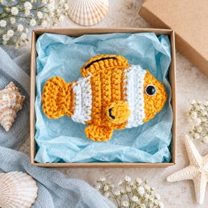 May include: A crocheted clownfish toy with orange and white horizontal stripes, a black-rimmed dorsal fin, and a single black plastic eye, nestled inside a square cardboard gift box lined with light blue tissue paper.