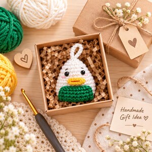 May include: A small, white, crocheted duck-shaped ornament with a yellow beak and a green base, nestled inside a square cardboard gift box filled with brown paper crinkle filler. The ornament features a small loop at the top for hanging.