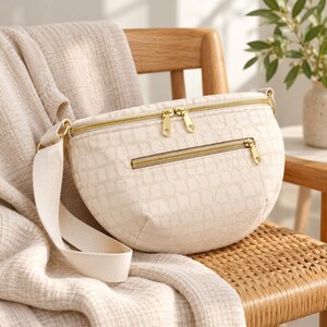 May include: A cream-colored crescent-shaped crossbody bag featuring a subtle embossed grid pattern, gold-tone metal zippers, and a wide matching fabric strap. The bag includes a main zippered compartment and a smaller front zippered pocket.