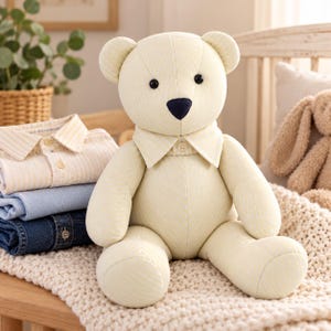 May include: A stuffed teddy bear made of pale yellow fabric with a subtle white herringbone pattern. The bear features a dark navy blue nose, black bead eyes, and a fabric collar with a single button at the center of the neck.