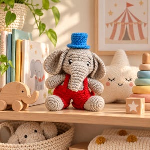 May include: A crocheted gray elephant toy wearing bright red overalls and a small blue top hat. The elephant has large ears, a trunk, and embroidered eyes. It is positioned among various children's toys, including books and a star-shaped pillow.