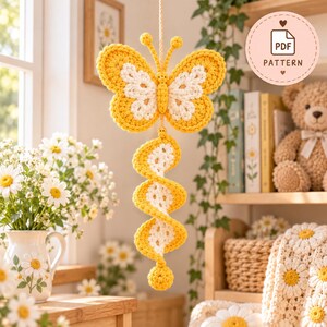 Puede incluir: Un m&oacute;vil de mariposa de ganchillo colgante con alas de hilo amarillo y blanco y una cola en espiral que termina en una peque&ntilde;a esfera amarilla. La parte superior incluye una insignia circular con el texto PDF PATTERN a la derecha.