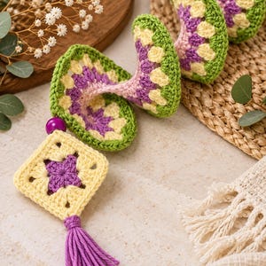 K&ouml;nnte beinhalten: Eine spiralf&ouml;rmige H&auml;kel-H&auml;ngedekoration bestehend aus einer Reihe verbundener Granny Squares in den Farben Hellgelb, Violett und Gr&uuml;n. Das St&uuml;ck ist mit einer violetten Perle und einer violetten Garnquaste am unteren Ende versehen.