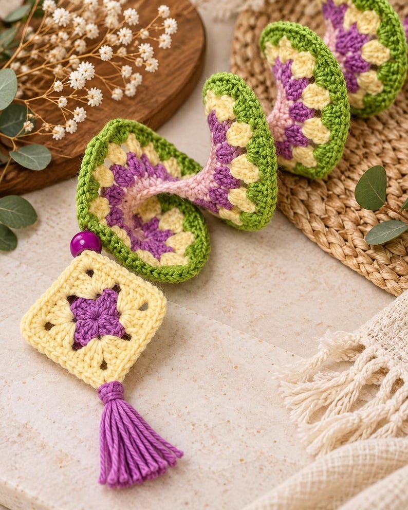 K&ouml;nnte beinhalten: Eine spiralf&ouml;rmige H&auml;kel-H&auml;ngedekoration bestehend aus einer Reihe verbundener Granny Squares in den Farben Hellgelb, Violett und Gr&uuml;n. Das St&uuml;ck ist mit einer violetten Perle und einer violetten Garnquaste am unteren Ende versehen.