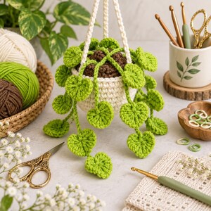 May include: A crocheted hanging plant with bright green heart-shaped leaves and brown soil texture in a cream-colored pot. The plant is suspended by a thick white crocheted cord, showcasing intricate yarn work and detailed leaf patterns.