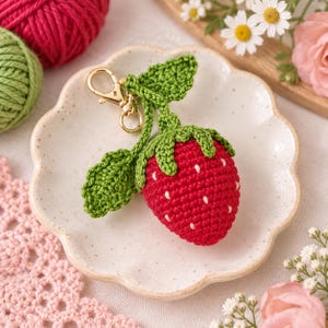 May include: A crocheted strawberry keychain featuring a bright red berry body with white embroidered seed details, a green crocheted stem with two leaves, and a gold-toned metal lobster clasp attached to the top for secure fastening.