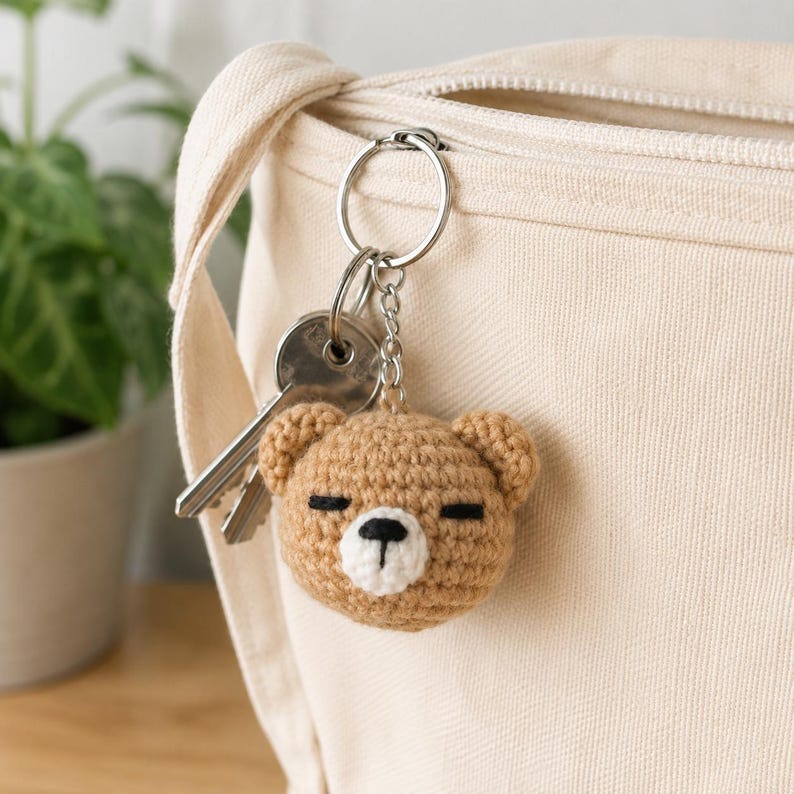 May include: A light brown crocheted bear head keyring with a white muzzle, black embroidered eyes, and a black nose. The bear is attached to a silver metal split ring and chain, which also holds a set of two silver metal keys.