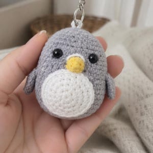 May include: A small, round, gray crocheted penguin keychain featuring a white circular belly patch, a yellow beak, and two black plastic bead eyes. The penguin is attached to a silver-toned metal keyring loop at the top of its head.