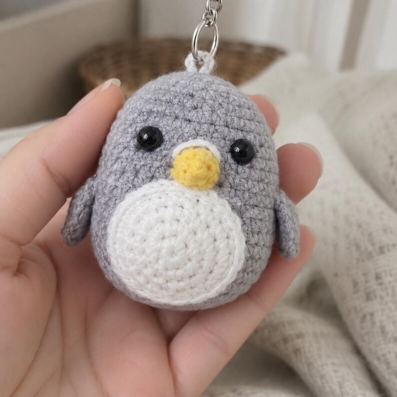 May include: A small, round, gray crocheted penguin keychain featuring a white circular belly patch, a yellow beak, and two black plastic bead eyes. The penguin is attached to a silver-toned metal keyring loop at the top of its head.