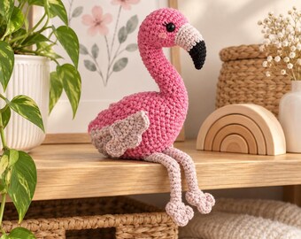 May include: A crocheted flamingo plush toy featuring a bright pink body, light pink legs, and a white beak with a black tip. The stuffed animal has a long curved neck, small black eyes, and textured wings, designed to sit upright on a flat surface.
