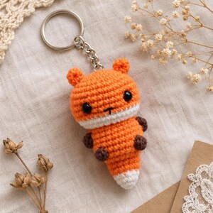 May include: A small, handmade crochet fox keychain featuring a bright orange head and body, white muzzle and tail tip, and dark brown paws. The fox has two round black plastic eyes and is attached to a silver-colored metal split ring by a short chain.
