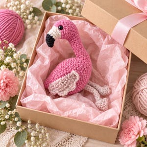 May include: A crocheted flamingo toy with a bright pink body, a white face, and a black beak. The flamingo has light pink wings and legs, and it is nestled inside a cardboard gift box lined with pink tissue paper, presented as a gift item.