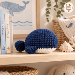 May include: A dark blue and white crocheted whale plush toy with a rounded body, a small tail, and a single black bead eye. The whale is positioned on a wooden shelf next to a stack of books and a woven basket containing a patterned fabric.