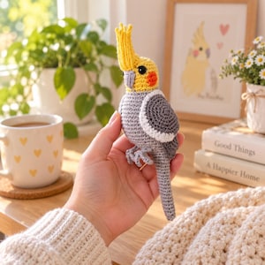 May include: A crocheted cockatiel bird toy featuring a yellow head with a crest, orange cheek patches, a gray body with white wing accents, and a long gray tail. The bird has black bead eyes and a small gray beak, crafted with detailed yarn stitches.