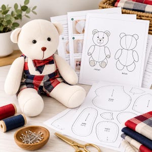 May include: A cream-colored plush teddy bear wearing red and blue plaid overalls and a matching bowtie, accompanied by printed sewing pattern instructions, fabric swatches, spools of thread, a bowl of pins, and gold scissors.