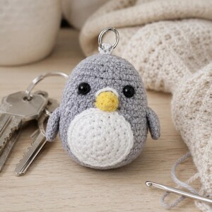 May include: A small, round, gray crocheted penguin keychain with a white belly, a yellow beak, and two black bead eyes. The penguin has a metal ring attached to the top for keys, and it is positioned next to a set of silver metal keys.
