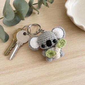 May include: A gray crocheted koala keychain featuring white inner ears, a black nose, and black eyes, holding a small light green crocheted leaf. The koala is attached to a silver metal split ring that holds two standard metal keys.