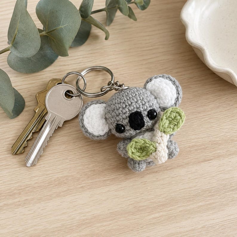 May include: A gray crocheted koala keychain featuring white inner ears, a black nose, and black eyes, holding a small light green crocheted leaf. The koala is attached to a silver metal split ring that holds two standard metal keys.