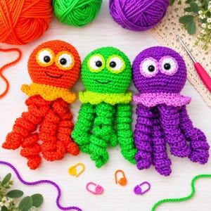 May include: Three crocheted octopus toys in bright orange, lime green, and purple. Each octopus features large white eyes with black pupils, a stitched smile, and eight curly tentacles. Small plastic stitch markers are scattered nearby.