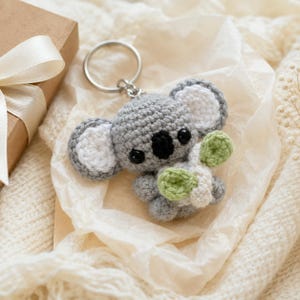 May include: A gray and white crocheted koala keychain featuring a round head, large ears with white centers, black plastic eyes, a black nose, and small limbs holding a light green and white crocheted leaf accessory attached to a silver metal ring.