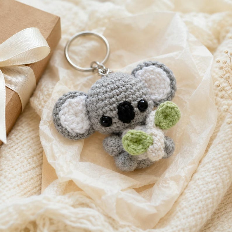 May include: A gray and white crocheted koala keychain featuring a round head, large ears with white centers, black plastic eyes, a black nose, and small limbs holding a light green and white crocheted leaf accessory attached to a silver metal ring.