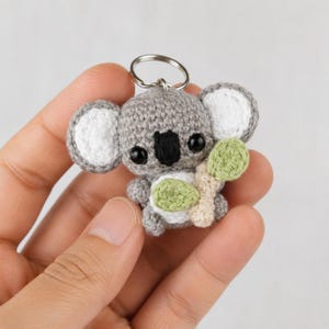 Peut inclure: Un petit porte-clés koala en crochet fait main avec un corps gris, des oreilles intérieures blanches et un nez noir. Le koala a des yeux en plastique noir et tient une branche d'eucalyptus crochetée vert clair et beige. Un anneau en métal argenté est fixé au sommet.