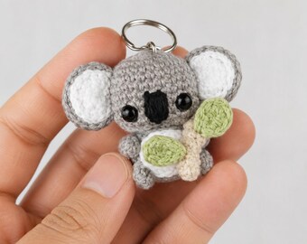 May include: A small, handmade crochet koala keychain featuring a grey body, white inner ears, and a black nose. The koala has black plastic eyes and is holding a light green and beige crocheted eucalyptus branch. A silver metal ring is attached to the top.