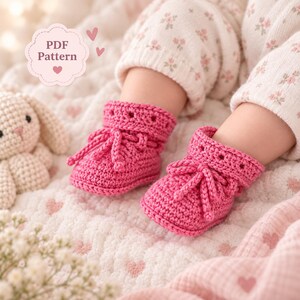 Op de afbeelding: Een paar felroze gehaakte babyslofjes met een omgeslagen boord met oogjes en een decoratief trekkoord. De slofjes worden gedragen aan voeten met een broek met bloemenprint, met een PDF Pattern label zichtbaar in de hoek van de afbeelding.