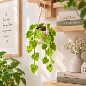 May include: A crocheted hanging plant featuring a cream-colored pot and vibrant green heart-shaped leaves on trailing vines. The plant is suspended by a cream-colored cord, showcasing detailed yarn textures and a realistic trailing vine appearance.