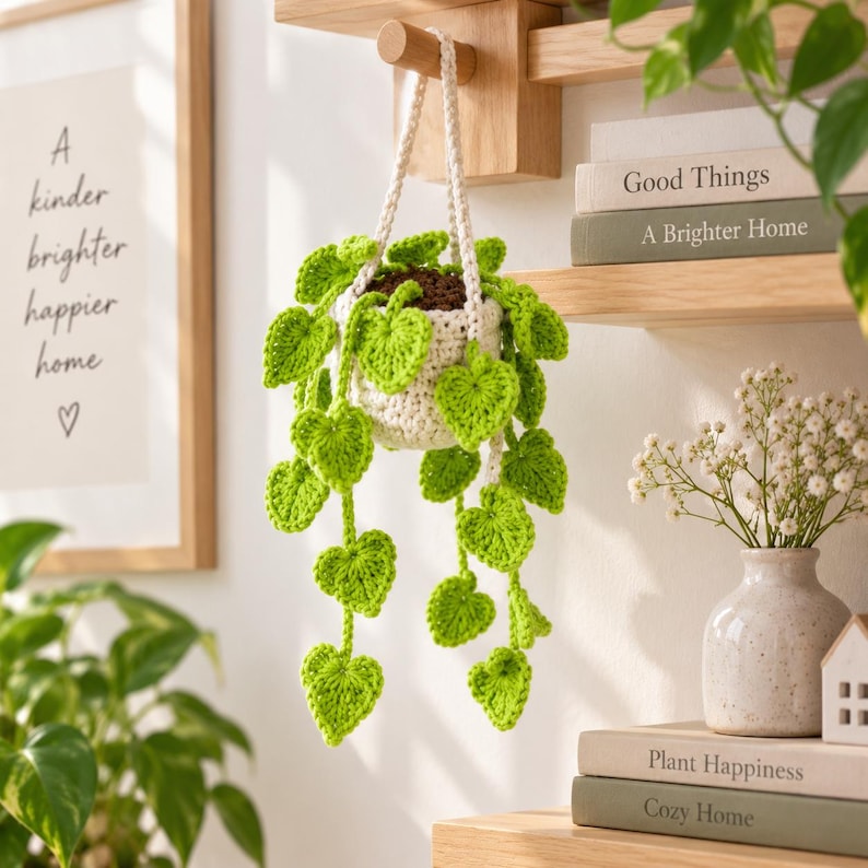 May include: A crocheted hanging plant featuring a cream-colored pot and vibrant green heart-shaped leaves on trailing vines. The plant is suspended by a cream-colored cord, showcasing detailed yarn textures and a realistic trailing vine appearance.