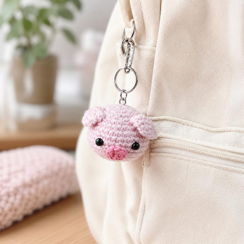 May include: A small, round, light pink crocheted pig keychain featuring two small ears, a darker pink snout, and two black bead eyes. The pig is attached to a silver-colored metal keyring and lobster clasp, which is clipped onto a cream-colored backpack.