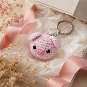May include: A round, light pink crocheted pig keychain featuring two small ears, a darker pink snout, and two black bead eyes. The pig is attached to a silver-toned metal split ring by a short chain, presented inside a gift box with tissue paper.