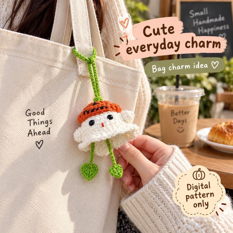 May include: A cream-colored canvas tote bag with the text Good Things Ahead and a heart symbol, featuring a crocheted ghost charm with an orange pumpkin hat, black eyes, and two green leaf-shaped dangling feet attached by a green yarn loop.