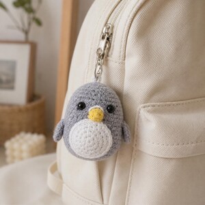 May include: A small, round, crocheted penguin keychain featuring a gray body, a white belly, and a yellow beak. The penguin has two black plastic eyes and is attached to a silver-toned metal keyring, which is clipped onto a cream-colored canvas backpack.