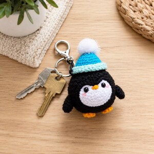 May include: A crocheted penguin keychain featuring a black and white body, orange feet, and a bright blue knit hat topped with a white pom-pom. The penguin is attached to a silver metal keyring that holds two standard silver-colored metal keys.