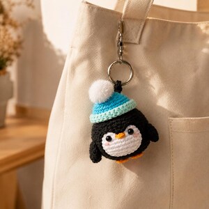 May include: A crocheted penguin keychain featuring a black and white body, a bright blue knit hat with a white pom-pom, orange feet, and a small orange beak. The penguin is attached to a silver-toned metal split ring and a lobster clasp.