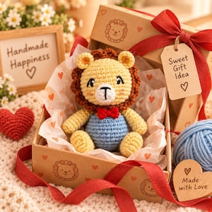 Puede incluir: Un le&oacute;n de peluche tejido a ganchillo con cara amarilla, melena marr&oacute;n y peto azul, con una peque&ntilde;a pajarita roja. El juguete est&aacute; dentro de una caja de regalo de cart&oacute;n con papel de seda de corazones, atado con una cinta roja y una etiqueta.