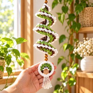 May include: A spiral-shaped crochet hanging ornament featuring alternating bands of green, brown, and white yarn. The piece is accented with wooden beads and terminates in a circular crochet motif with a small tassel at the bottom.