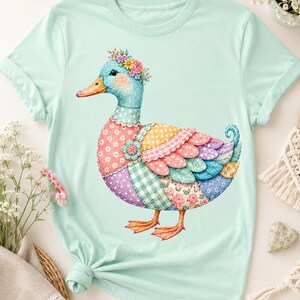 Peut inclure: Un t-shirt vert menthe clair avec un motif central représentant un canard portant une couronne de fleurs. Le corps du canard est composé de divers motifs en patchwork colorés, incluant des imprimés floraux, à pois et vichy dans des tons rose, bleu et jaune.